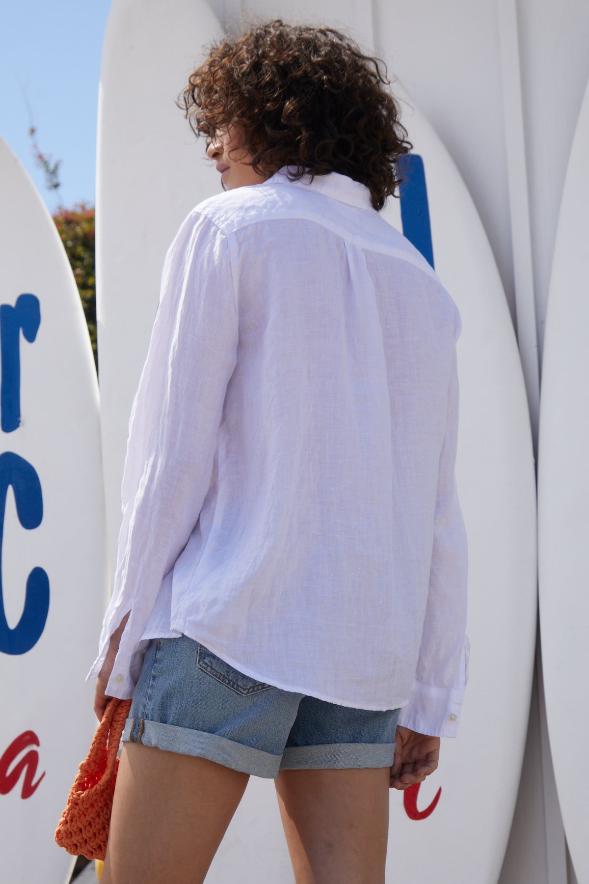 A person with curly hair is seen from the back, wearing a white long-sleeve NATALIA LINEN BUTTON-UP SHIRT by Velvet by Graham & Spencer and denim shorts with a relaxed silhouette, standing in front of surfboards.-35942448300225