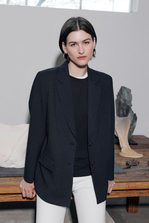 A person with short dark hair wears the FRESNO PINSTRIPE BLAZER by Velvet by Jenny Graham in an oversized fit, paired with a black shirt and white pants, standing indoors near a wooden table with decorative objects.