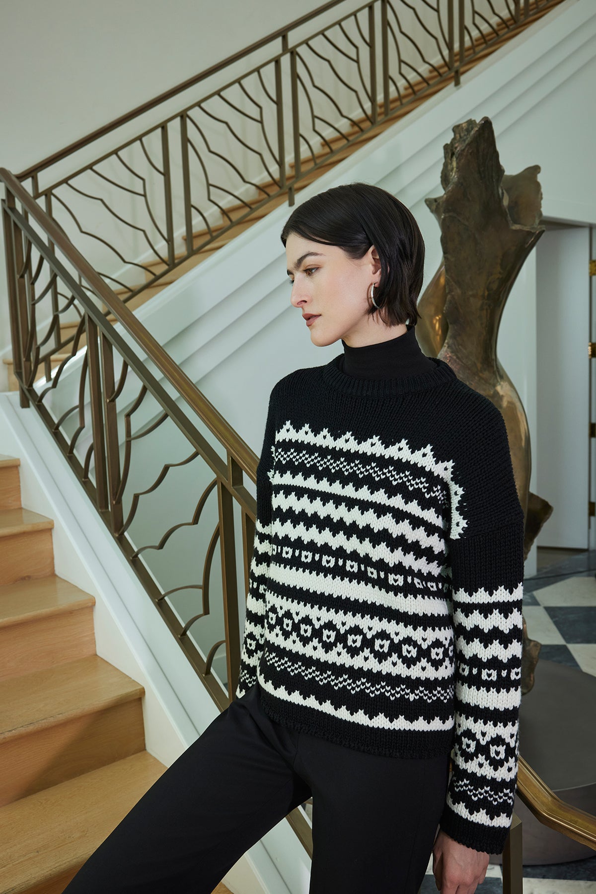 A person wearing the Velvet by Jenny Graham MAYWOOD SWEATER, featuring a relaxed fit and chunky wool blend knit with a fair isle pattern, stands by a staircase with a wooden railing.-38318585086145