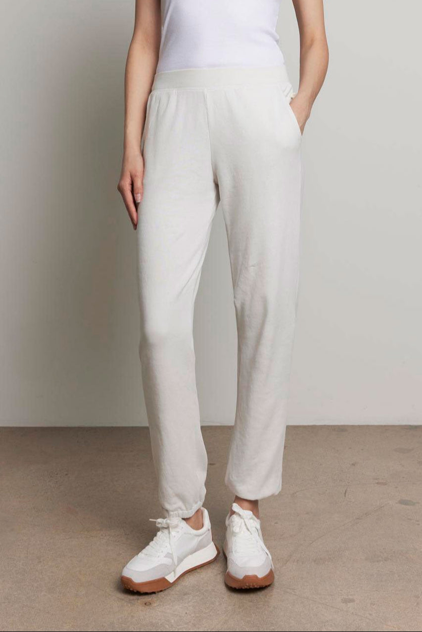 A person is standing against a plain background, dressed in a white tank top and the ZUMA SWEATPANT from Velvet by Jenny Graham, featuring off-white organic cotton material with an elastic waist and pockets, paired with white sneakers, looking at the camera with a neutral expression.-37817332629697