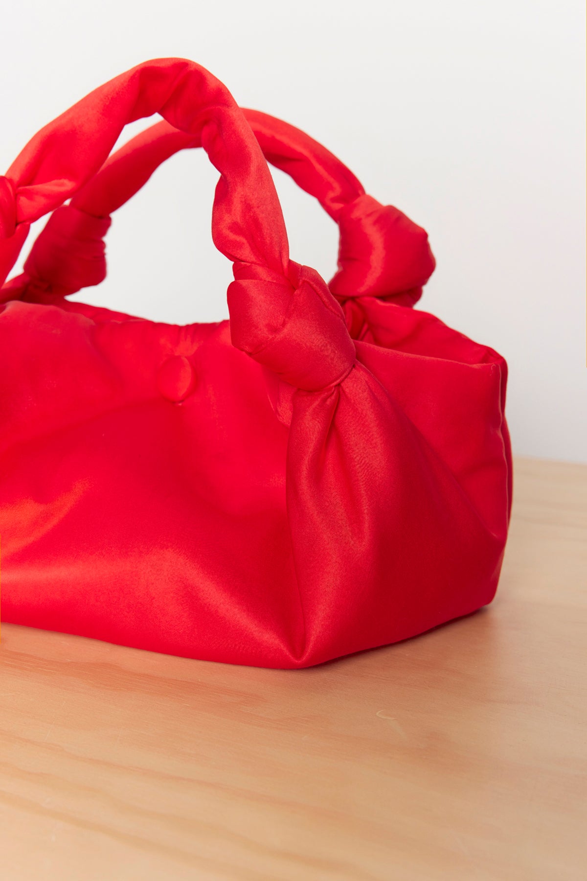 The ROBYN SATIN BAG by Velvet by Jenny Graham, featuring its vibrant red satin material and sophisticated knot details with coordinating knotted handles, is placed neatly on a wooden surface.-38318606418113