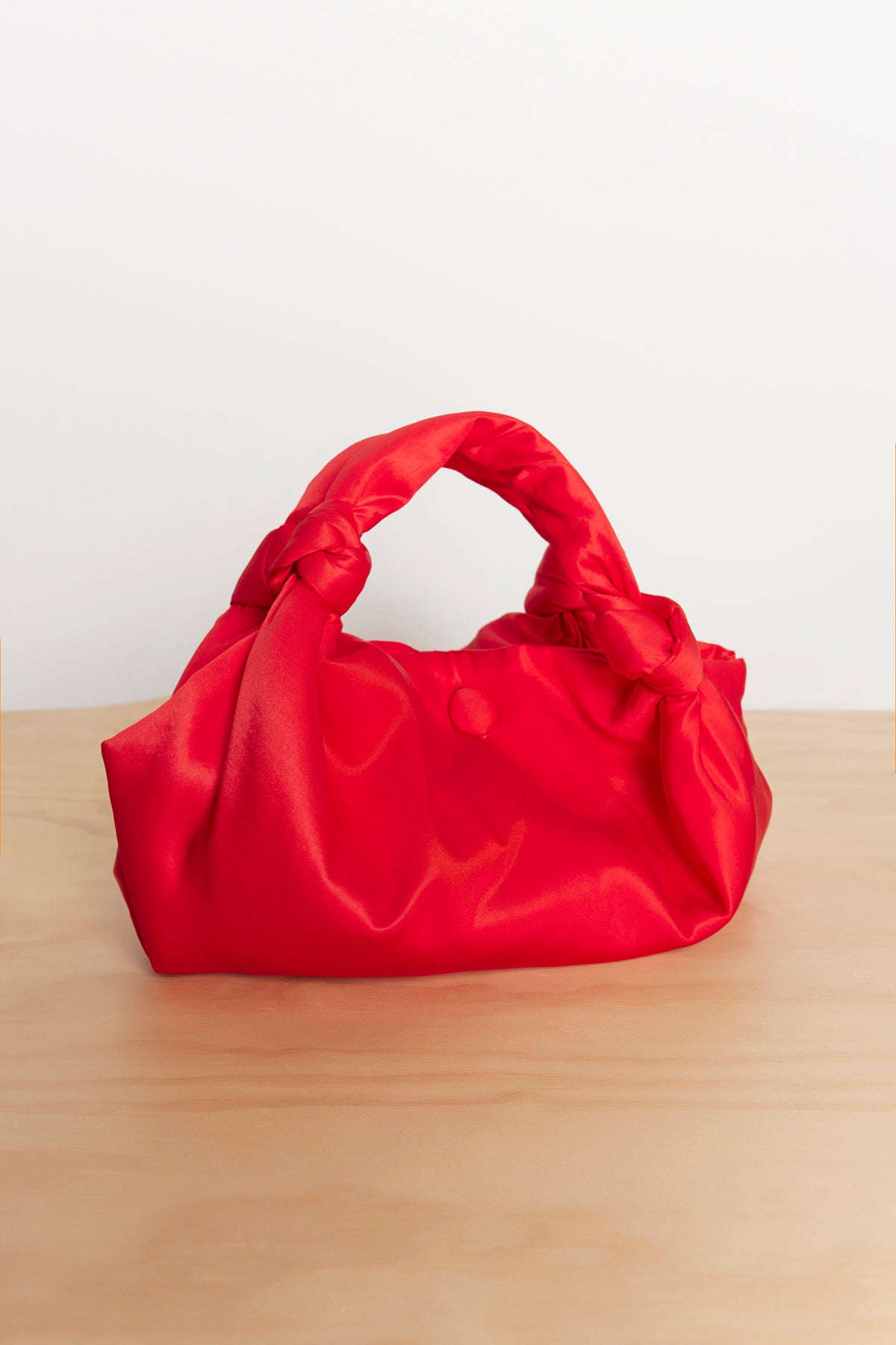 The ROBYN SATIN BAG by Velvet by Jenny Graham, featuring elegant knot details on the handle in a compact red satin design, is placed on a wooden surface.