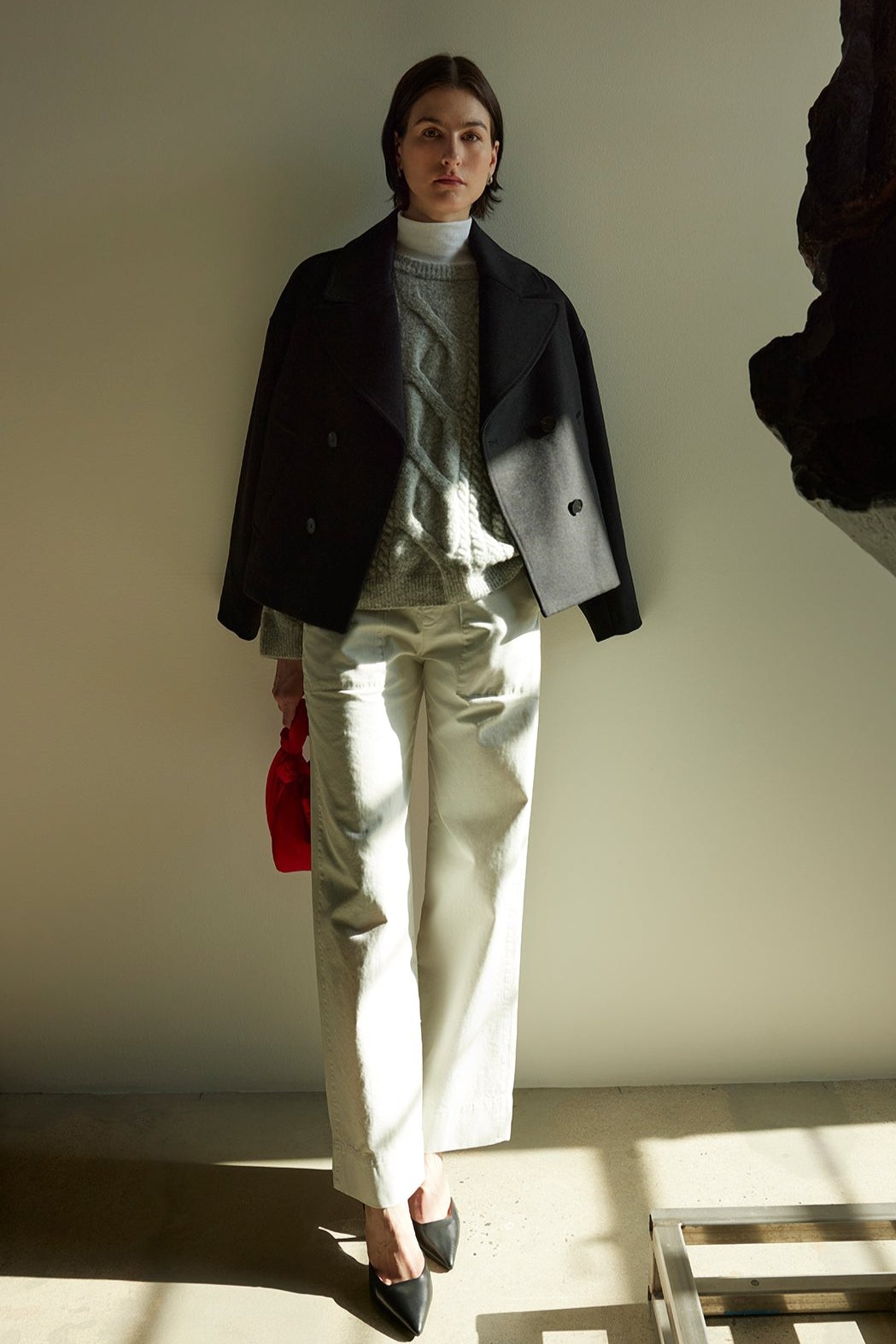 A person stands in a sunlit room wearing a dark jacket, light sweater, and the VENTURA PANT by Velvet by Jenny Graham, made of cotton twill. Black shoes ground the outfit while they hold a red bag next to a large abstract sculpture.-38318619263169