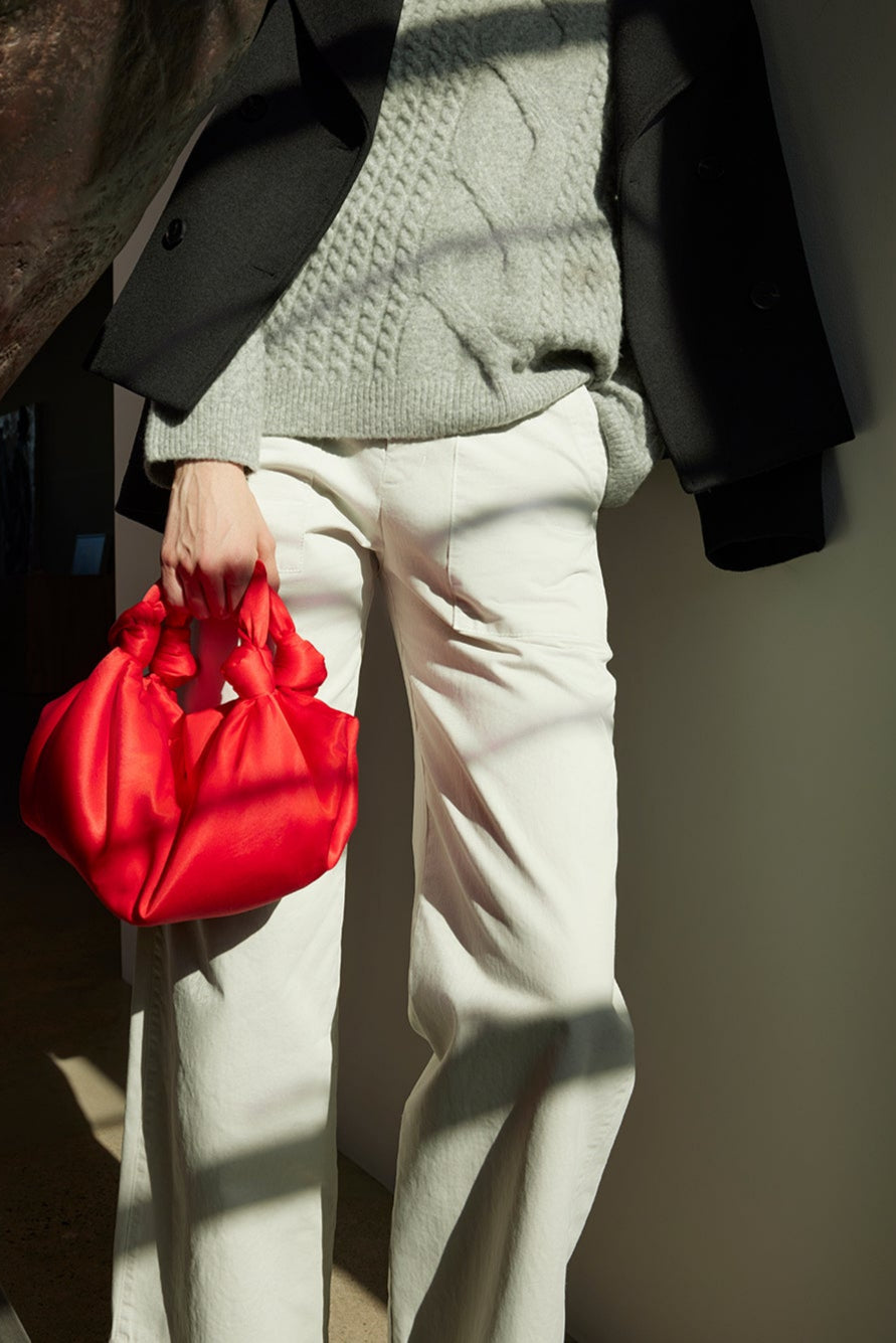 A person leans against a wall, dressed in a gray sweater, black coat, and white pants. They carry the ROBYN SATIN BAG from Velvet by Jenny Graham, which features elegant knot details. The bag's satin finish gleams softly against their shadow on the wall.
