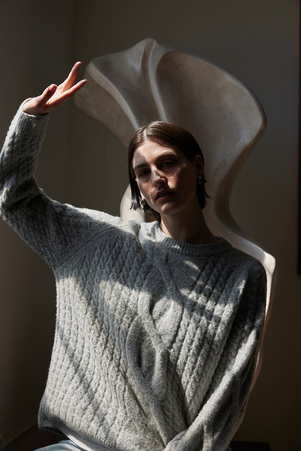 A person wearing the WILSHIRE SWEATER from Velvet by Jenny Graham, a cozy gray wool-yak blend sweater, poses with a peace sign in front of a large abstract sculpture.-38318578335937