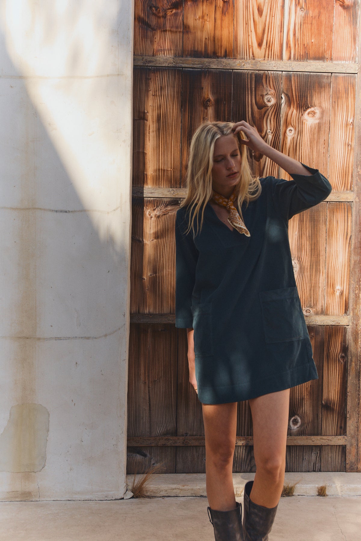 A woman with blonde hair stands before a wooden door, wearing the Velvet by Graham & Spencer ELKE CORDUROY DRESS, styled with boots and a scarf, one hand touching her head.-39744830537921