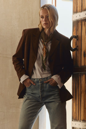 A person stands indoors by a wooden door, hands in pockets, wearing the SALIE FAUX SUEDE JACKET by Velvet by Graham & Spencer over a white shirt, paired with jeans and a patterned scarf, looking forward.