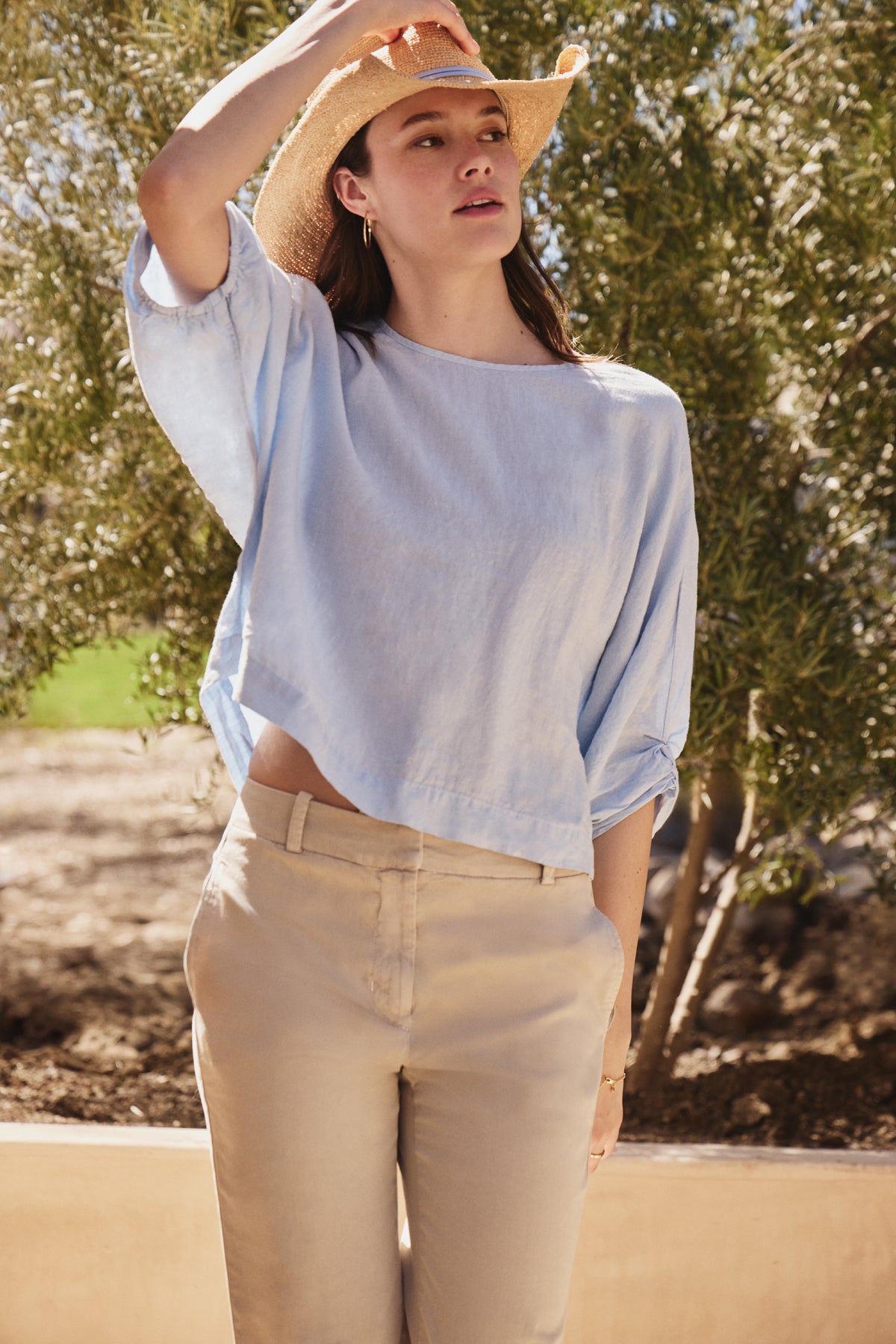 A person in a Velvet by Graham & Spencer CASS LINEN TOP in light blue and beige pants stands outdoors, donning a straw hat. Their hand rests on the brim, while the cocoon silhouette complements the lush greenery, evoking a serene summer day.-38928819585217