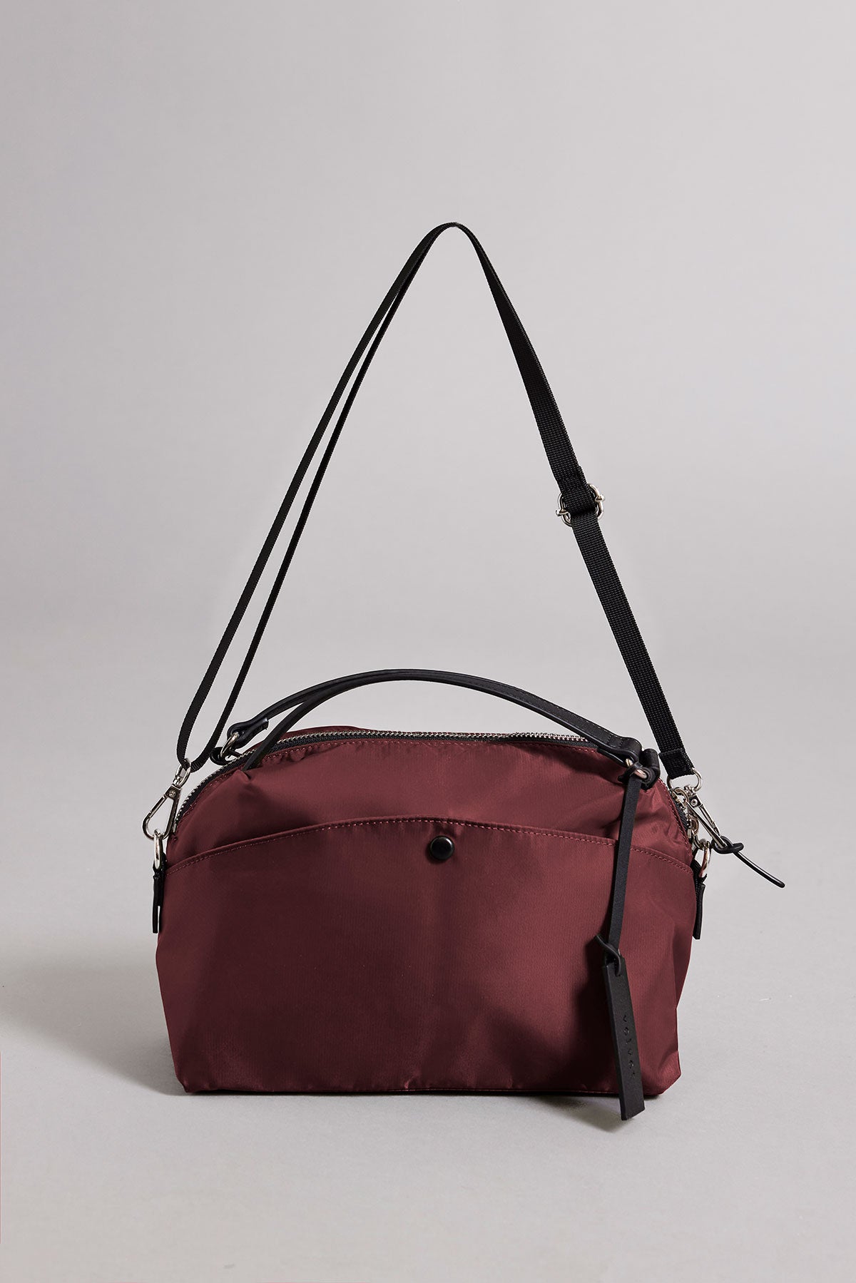 The NYLON CROSSBODY BAG by Velvet by Graham & Spencer is a maroon bag featuring a black adjustable strap, front pocket with button, top zipper closure, and black tag. Made of nylon, it's ideal for everyday use and shown on a plain light gray background.