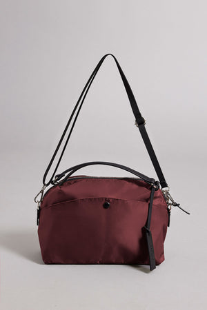 The NYLON CROSSBODY BAG by Velvet by Graham & Spencer is a maroon bag featuring a black adjustable strap, front pocket with button, top zipper closure, and black tag. Made of nylon, it's ideal for everyday use and shown on a plain light gray background.