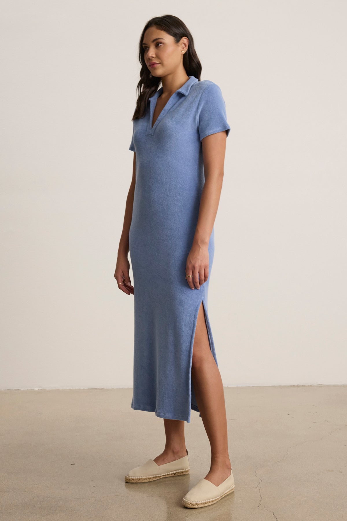 A woman stands indoors wearing the LIANNE POLO DRESS by Velvet by Graham & Spencer, a light blue, midi-length cotton loop terry dress with short sleeves, a collar, and side slit, paired with beige espadrilles.-39149167476929