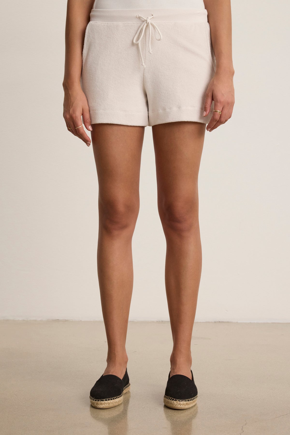 A person stands indoors wearing Velvet by Graham & Spencer's PEYTON SHORT—relaxed fit white cotton loop terry shorts with an elastic drawstring waist—paired with black espadrille shoes, hands at their sides.
