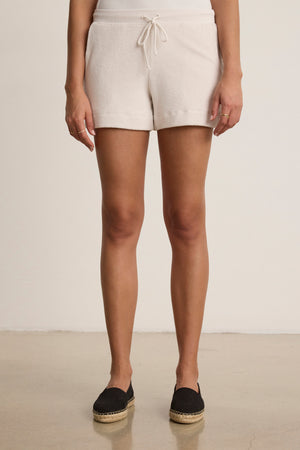 A person stands indoors wearing Velvet by Graham & Spencer's PEYTON SHORT—relaxed fit white cotton loop terry shorts with an elastic drawstring waist—paired with black espadrille shoes, hands at their sides.