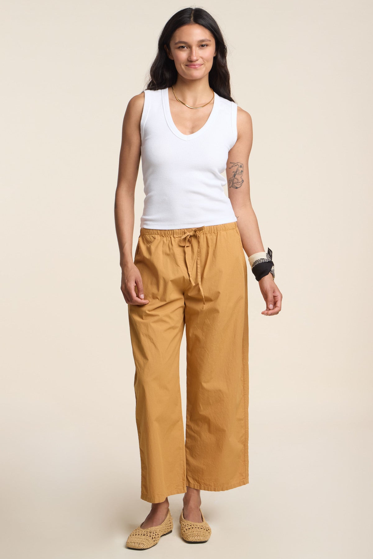 A person stands against a beige background wearing the DEMI TANK TOP by Velvet by Graham & Spencer, mustard drawstring pants, woven shoes, and bracelets.-41384065302721