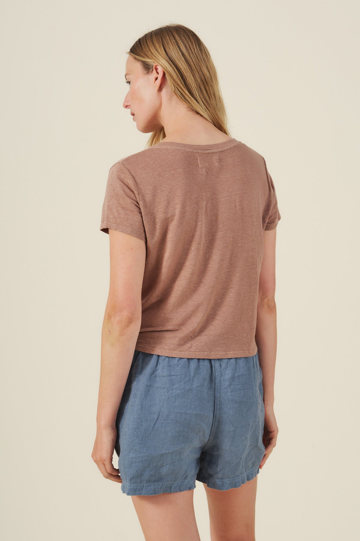 A person with long blonde hair is seen from behind, wearing the Velvet by Graham & Spencer AMBER TEE—a brown linen knit shirt with a ribbed V-neckline—paired with blue shorts against a plain background.-41526765158593