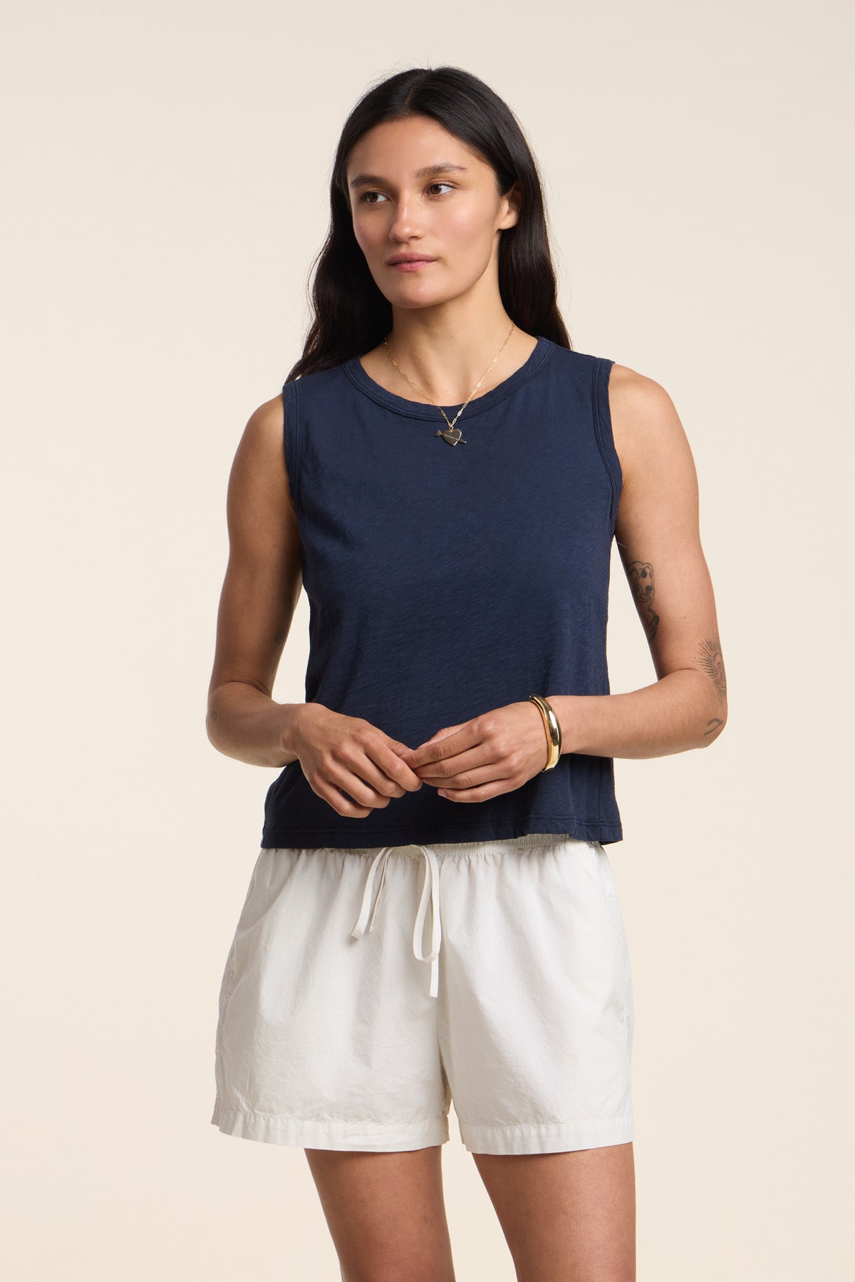 A woman with long dark hair wears the AMBRIA TANK TOP by Velvet by Graham & Spencer, styled in a boxy swing silhouette with white drawstring shorts, standing against a plain light background.-41526896328897