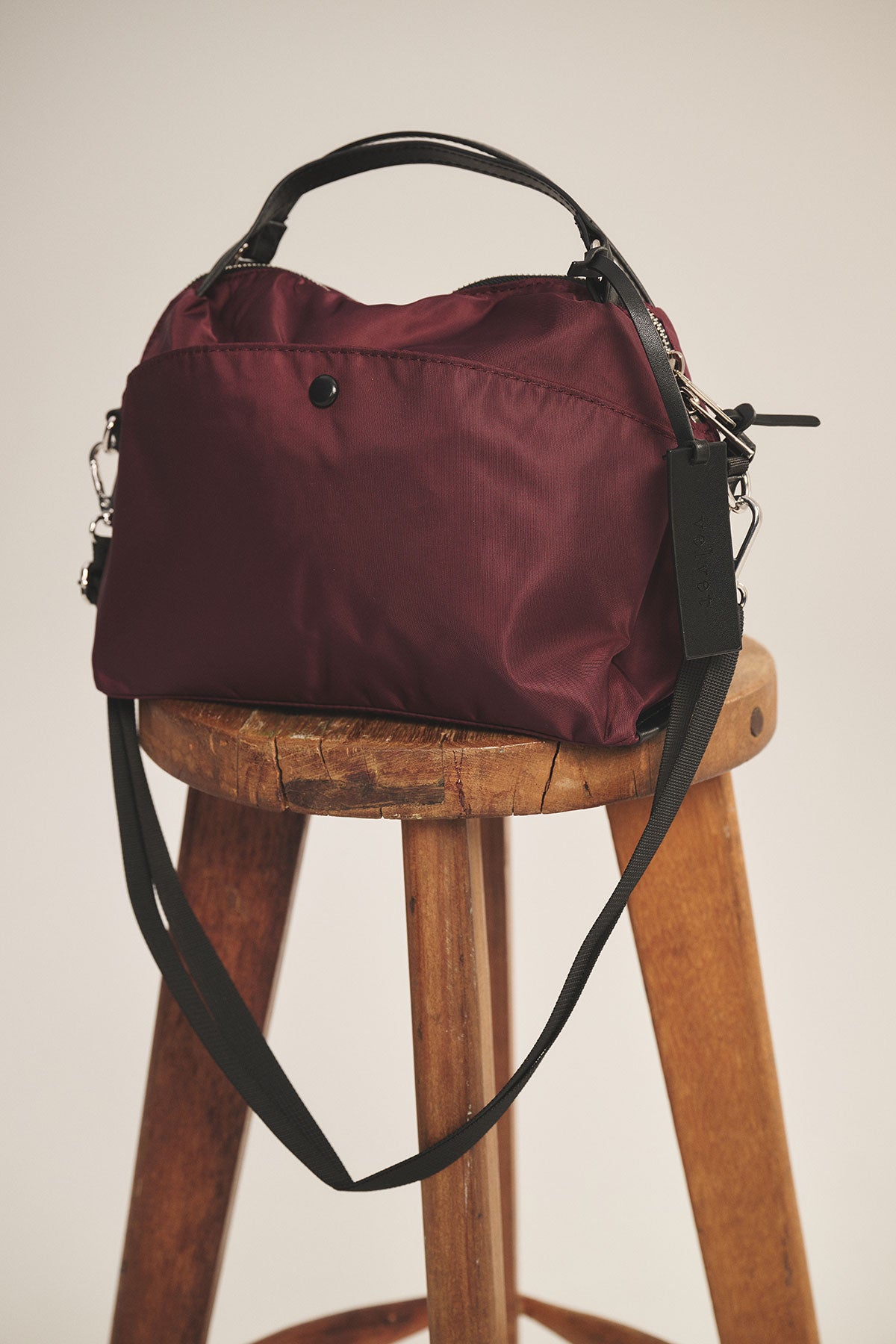 The NYLON CROSSBODY BAG by Velvet by Graham & Spencer in maroon, featuring black handles and a removable shoulder strap, is displayed on a wooden stool against a neutral background.
