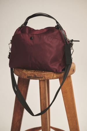 The NYLON CROSSBODY BAG by Velvet by Graham & Spencer in maroon, featuring black handles and a removable shoulder strap, is displayed on a wooden stool against a neutral background.