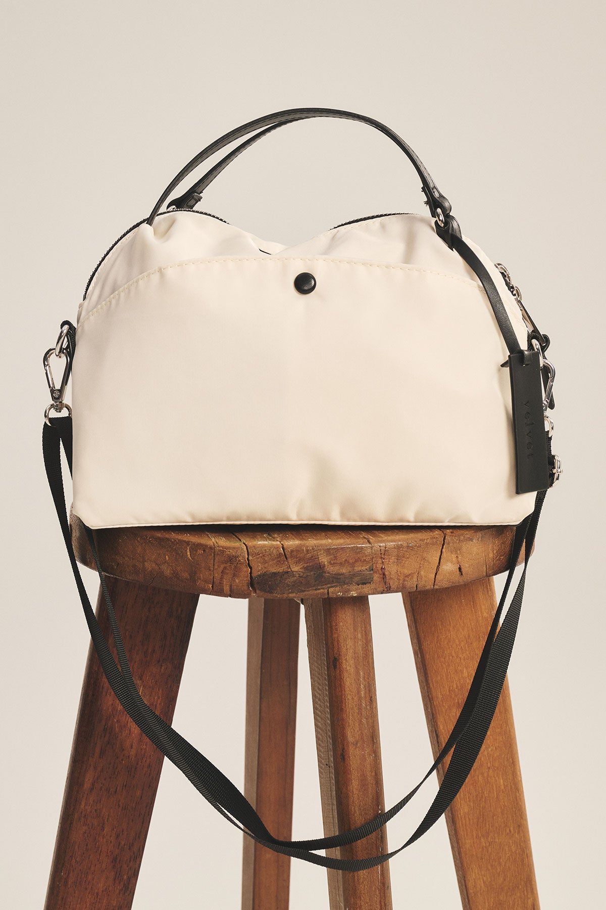 The NYLON CROSSBODY BAG by Velvet by Graham & Spencer, in cream with black handles and a removable shoulder strap, is displayed on a wooden stool against a plain background.