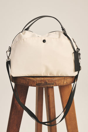 The NYLON CROSSBODY BAG by Velvet by Graham & Spencer, in cream with black handles and a removable shoulder strap, is displayed on a wooden stool against a plain background.