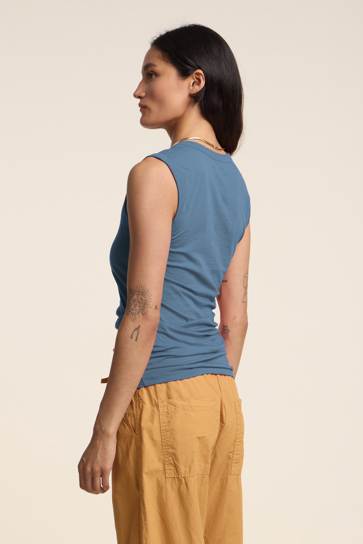 A person stands sideways against a plain background wearing the Velvet by Graham & Spencer ESTINA TANK TOP in blue, paired with tan pants and showcasing arm tattoos.-41384929034433
