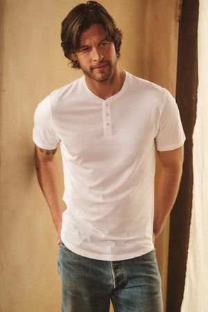 A man in a white FULTON HENLEY by Velvet by Graham & Spencer, paired with blue jeans and a curved hemline, stands indoors with his hands behind his back.