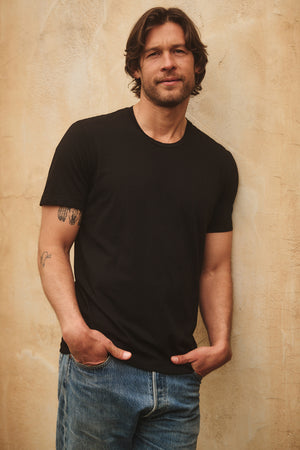 A man with shoulder-length hair in a HOWARD TEE by Velvet by Graham & Spencer, paired with jeans, leans against a textured wall with his hands in his pockets.