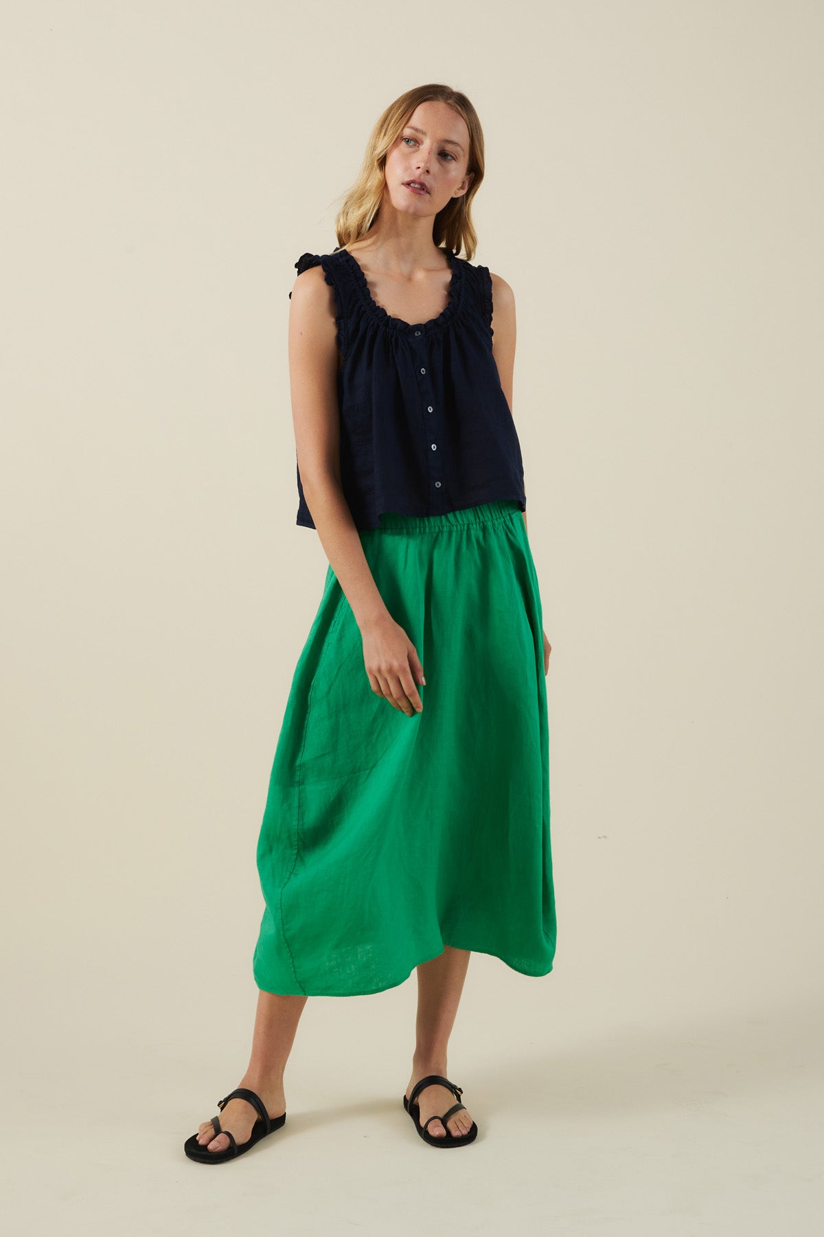 A woman stands against a plain background wearing a sleeveless navy blouse, black sandals, and the FAE LINEN SKIRT by Velvet by Graham & Spencer in an A-line green silhouette.-41527342596289