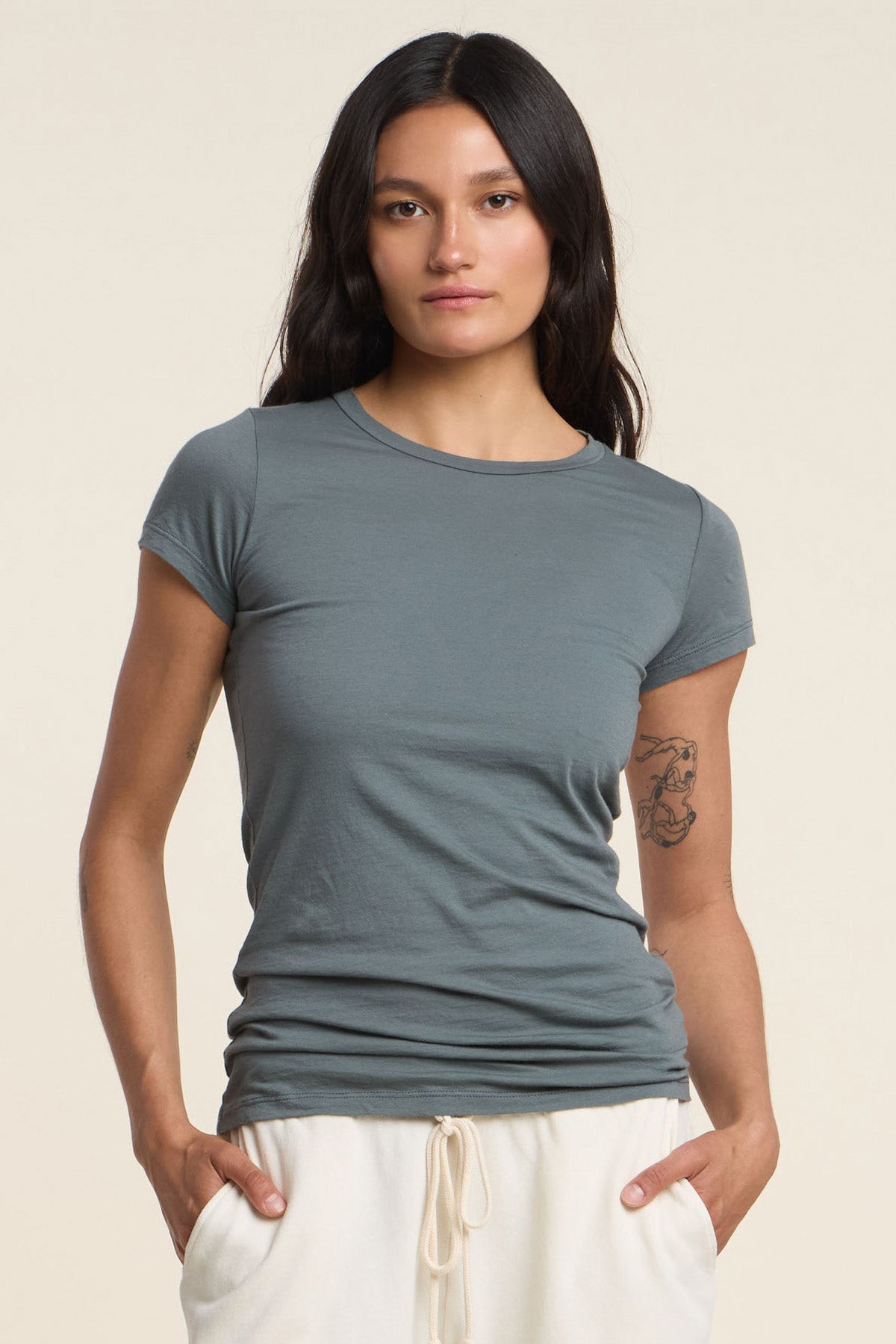 A person with long dark hair wears the JEMMA TEE by Velvet by Graham & Spencer and light drawstring pants, standing against a plain background with a tattoo visible on their left arm.-41374547968193