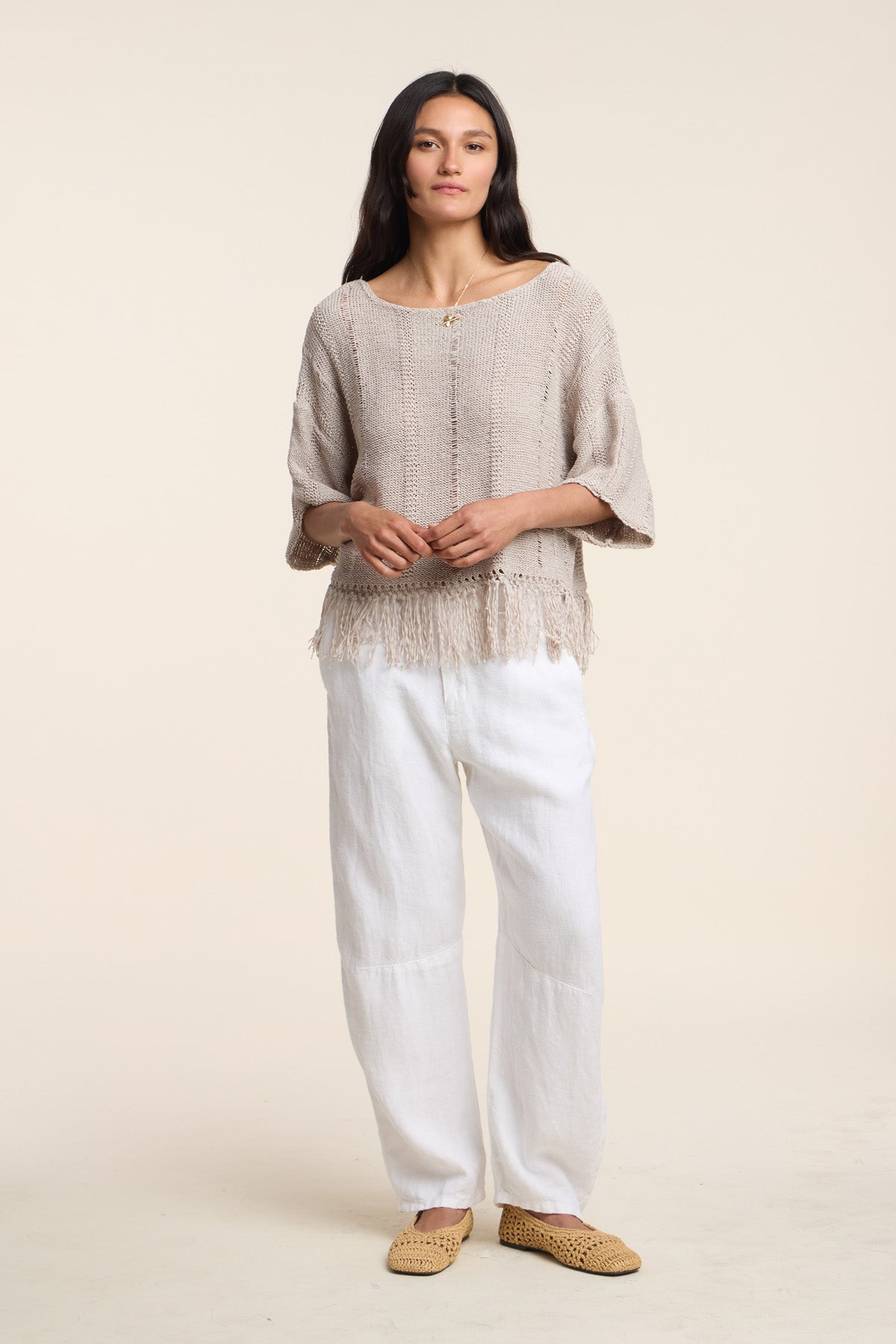 A woman faces forward against a plain light background, wearing the loose beige KATARINA SWEATER by Velvet by Graham & Spencer with a fringe hem, paired with white wide-leg pants and woven tan slip-on shoes.-41527058104513