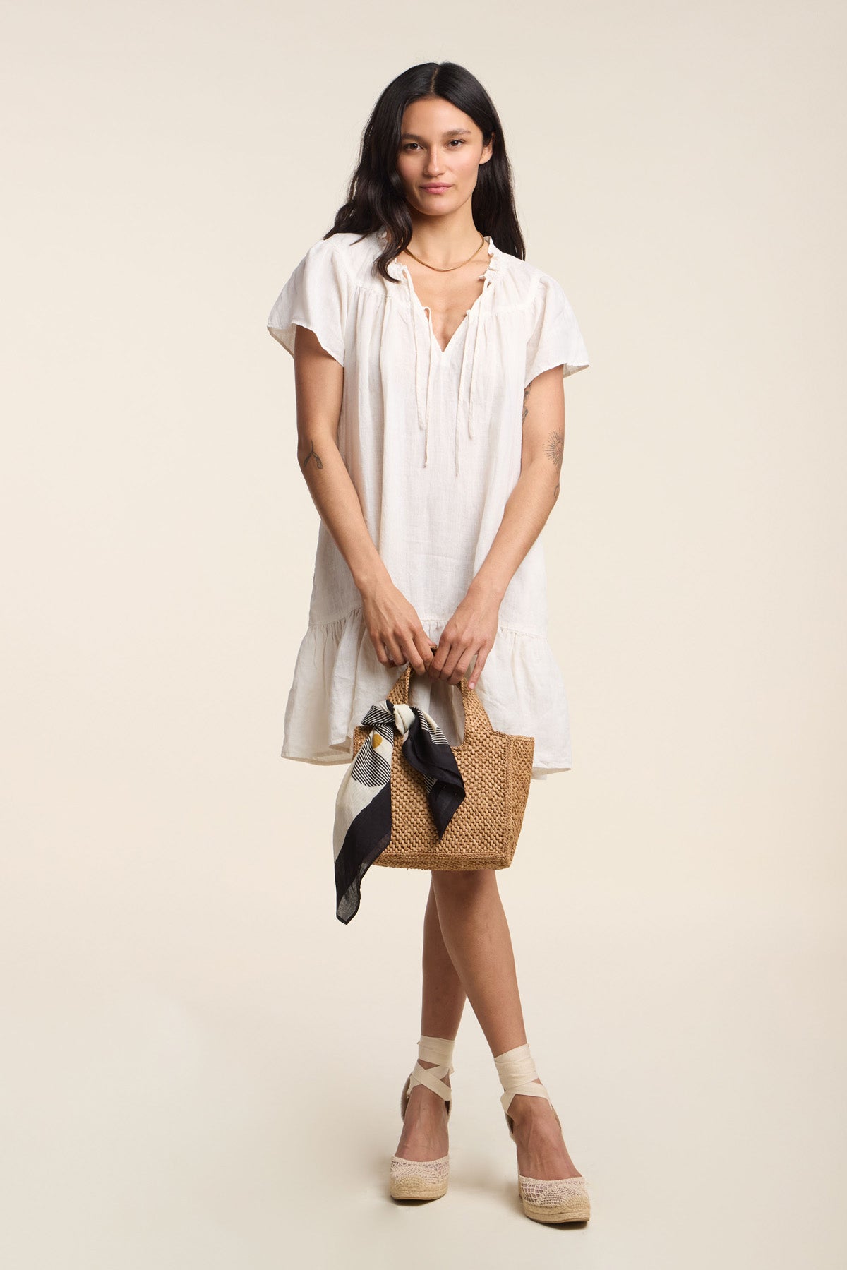 A woman wears the LIVVY LINEN DRESS by Velvet by Graham & Spencer, styled with a straw handbag and beige espadrilles, creating an effortless summer look against a plain background.-41371959230657