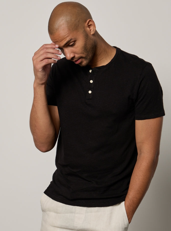 A man in a black short-sleeve shirt and light pants, with a shaved head, poses against a plain background. He is looking down and touching his forehead with one hand.