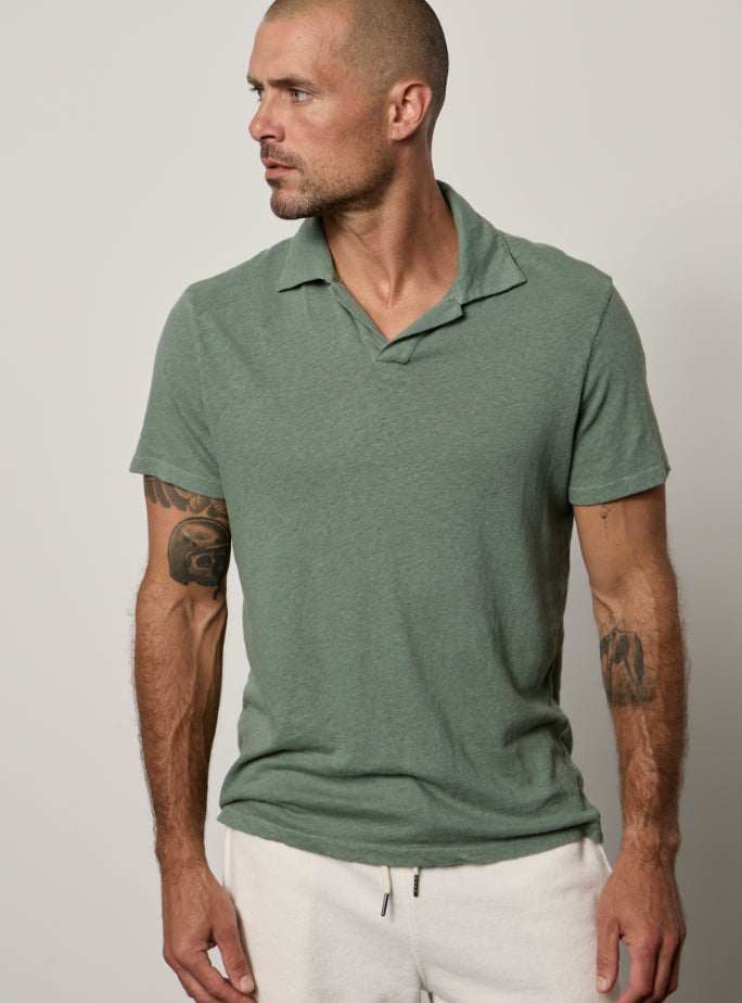 A man with a shaved head, trimmed beard, and tattoos on both arms, wearing a green polo shirt and white pants, looks to the side in front of a plain background.