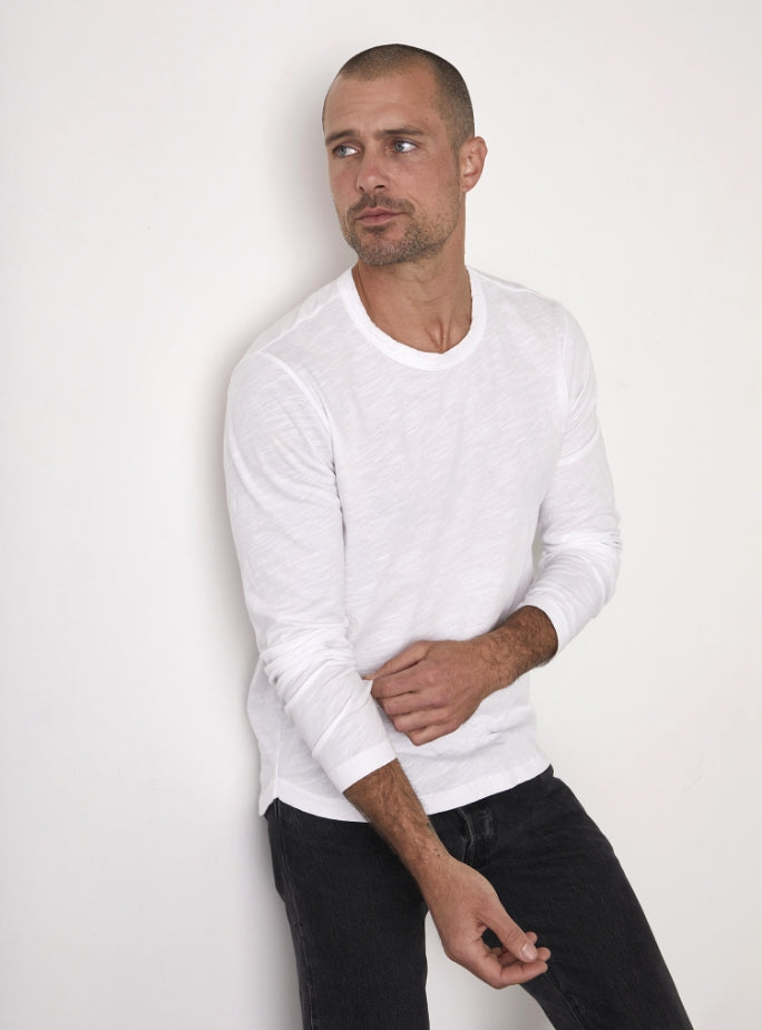 A man with closely cropped hair and a short beard is wearing a white long-sleeve shirt and dark pants, standing against a plain white background, looking to his left.