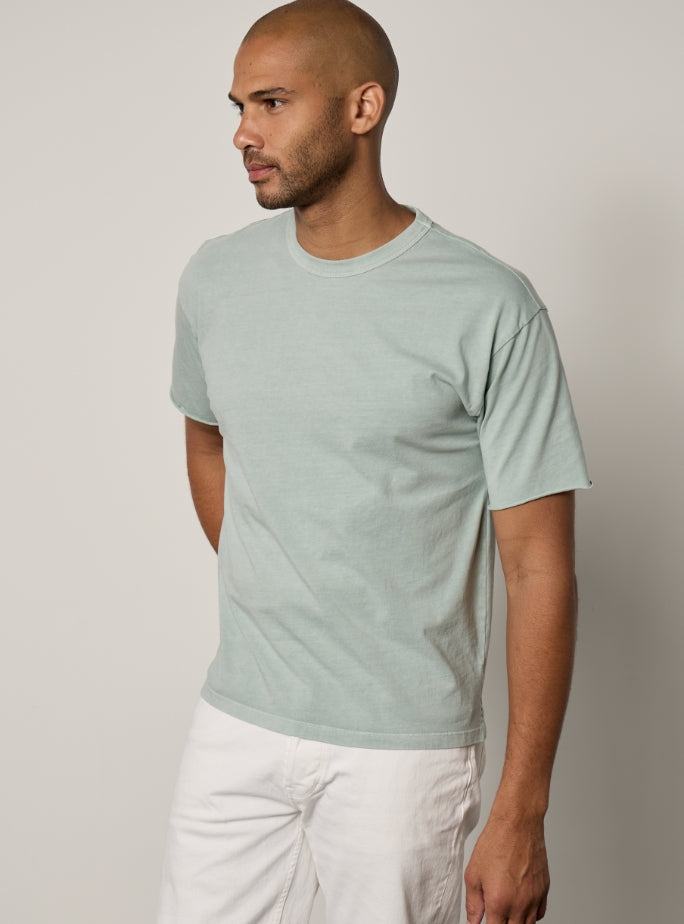A person with a shaved head and short beard is wearing a light green T-shirt and light gray pants, standing against a plain background, facing slightly to the right.