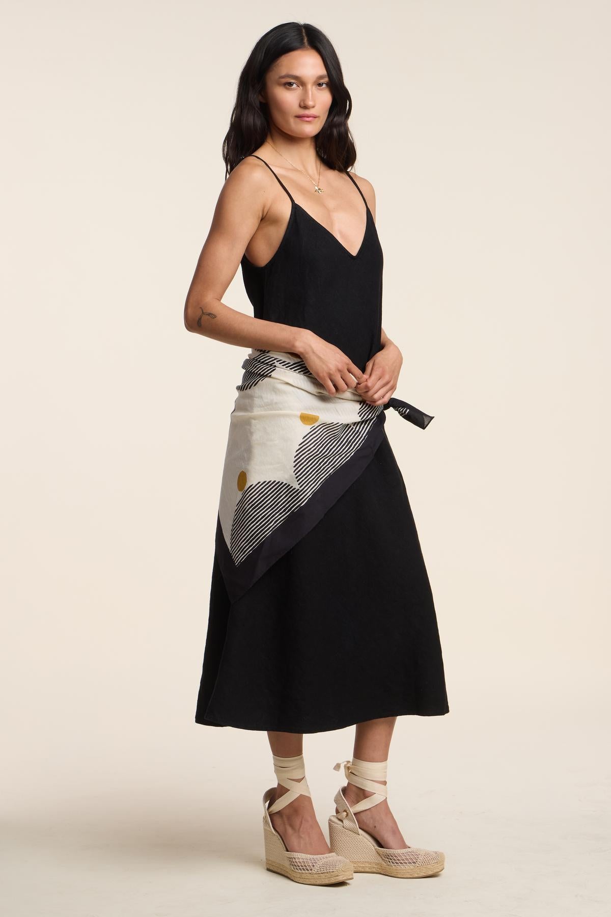 A woman stands against a beige background, wearing a black sleeveless midi dress with wedge espadrilles and the SOLI SCARF by Velvet by Graham & Spencer tied around her waist.-41393469358273