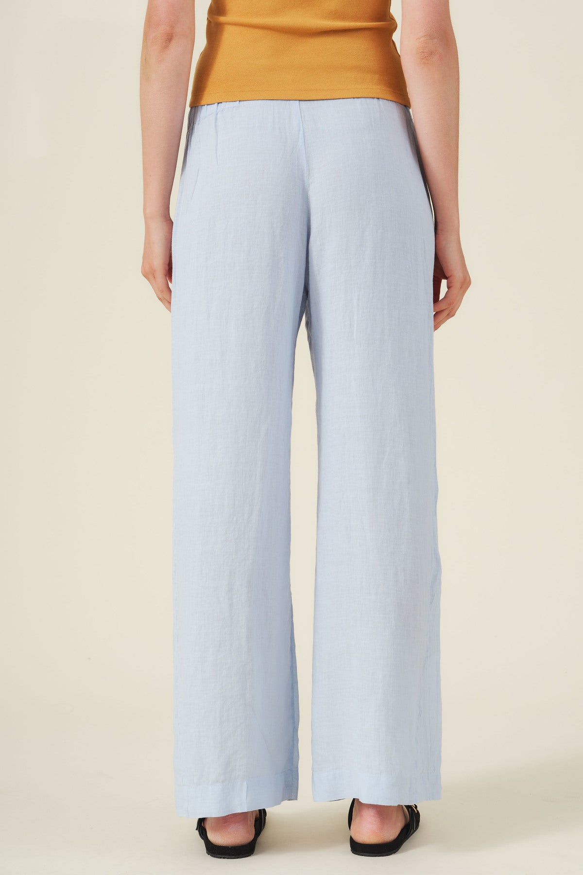 Person facing away wears Velvet by Graham & Spencer's STEPHANIE LINEN PANT in light blue with an elastic waist, paired with a mustard sleeveless top and black shoes against a neutral background.-41384915173569