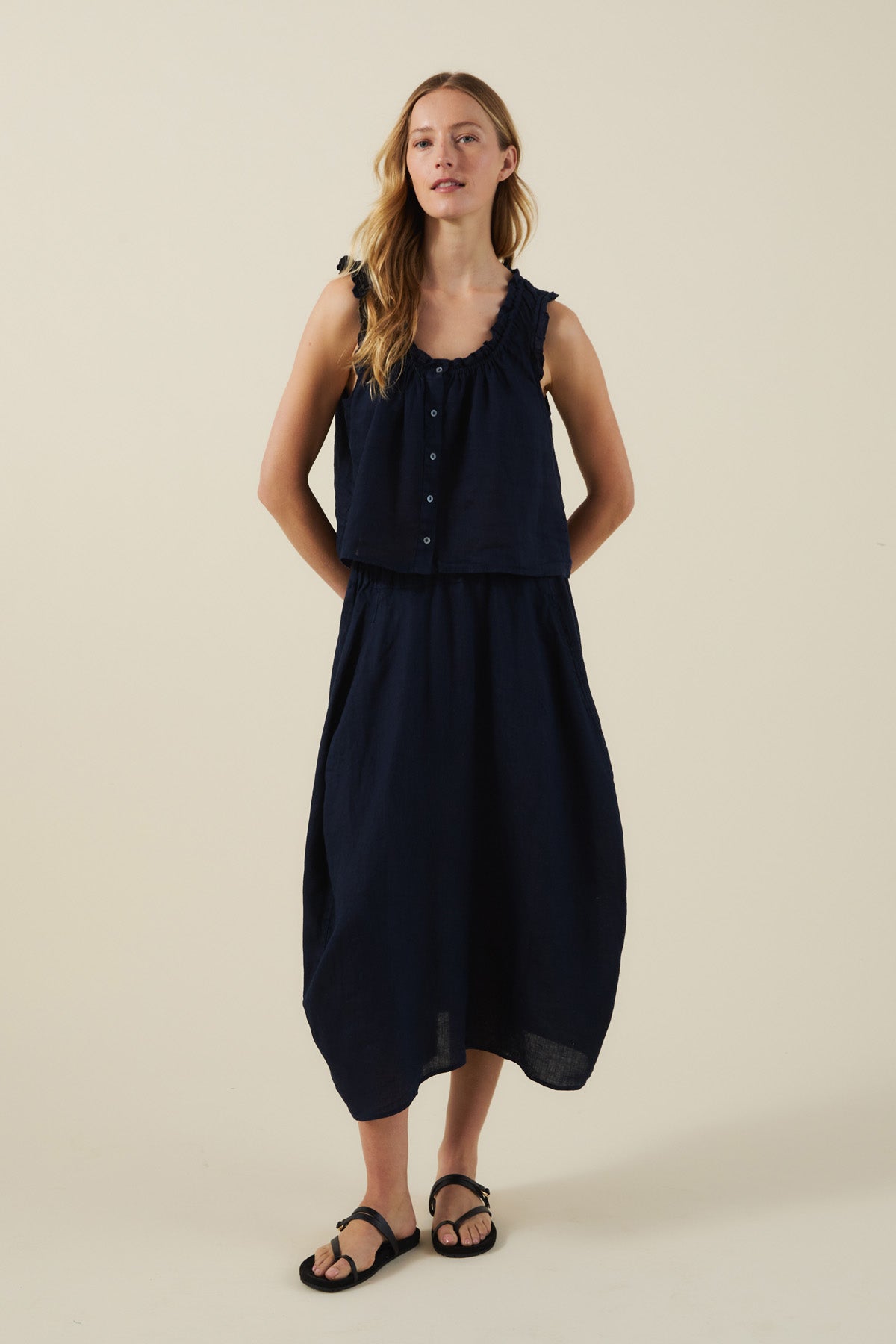 A woman stands against a plain background wearing the TANA LINEN TOP by Velvet by Graham & Spencer with a matching navy skirt and black sandals.-41527521116353