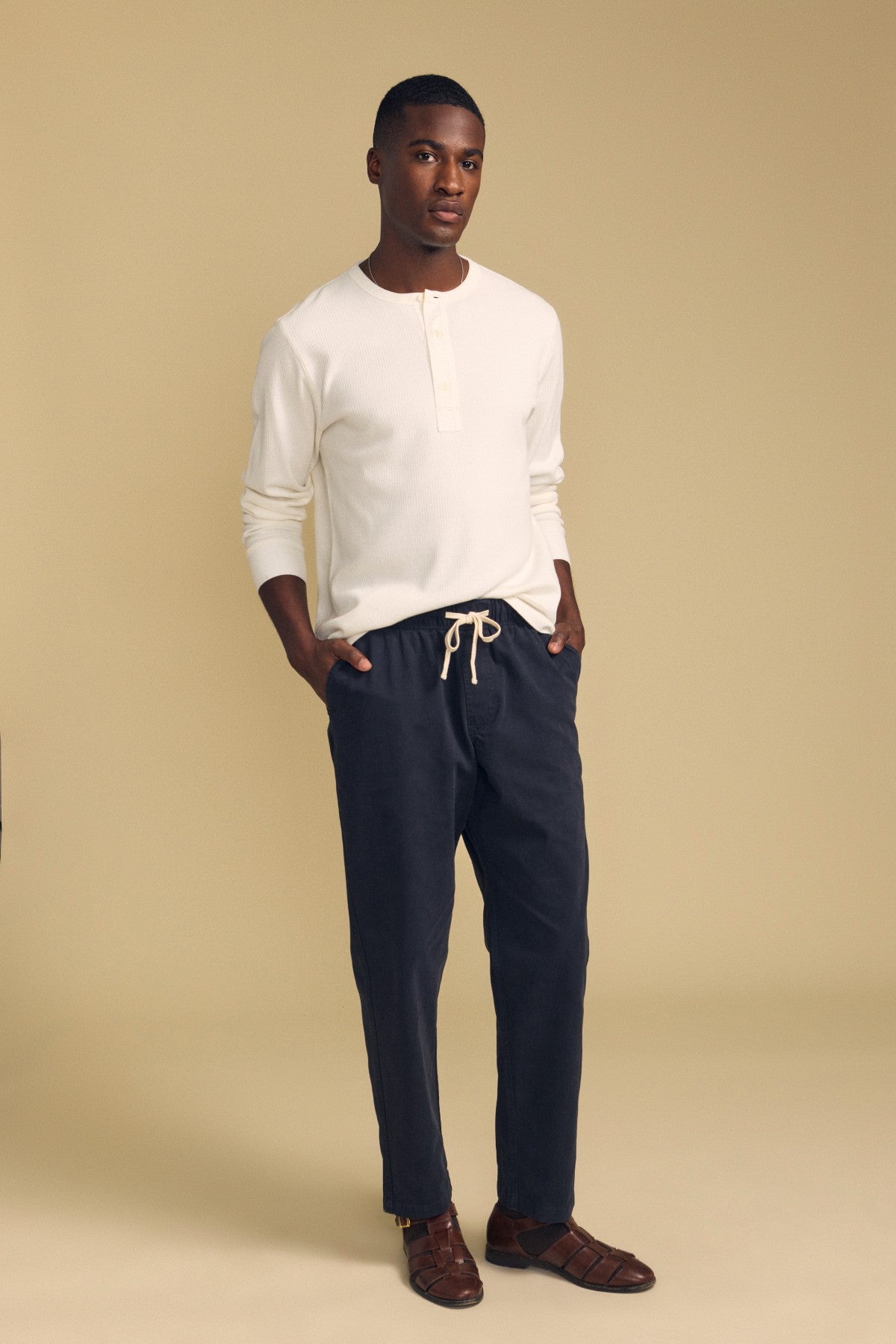 A man stands against a beige background, wearing the Velvet by Graham & Spencer CHANCE THERMAL HENLEY in white, paired with dark drawstring pants and brown sandals, with his hands in his pockets.-41859845226689