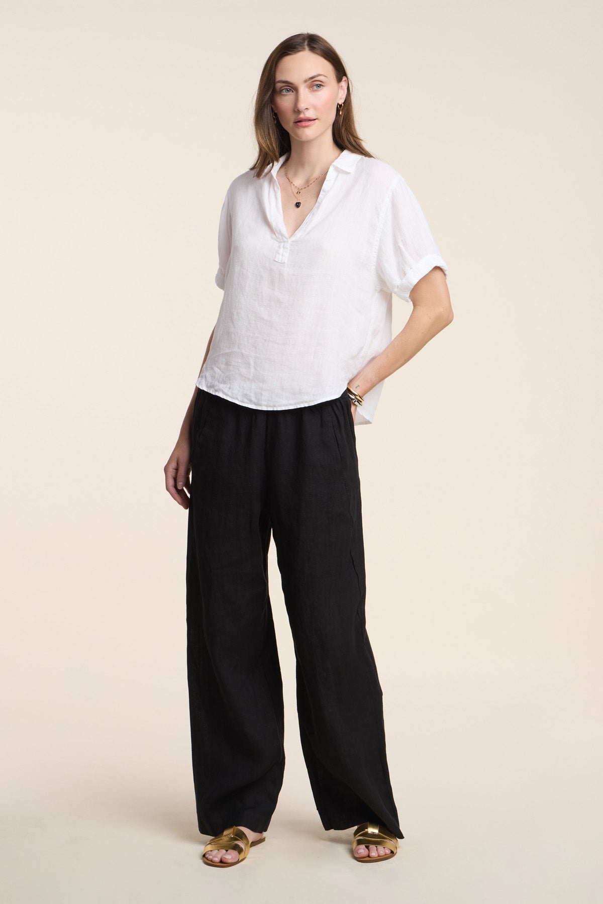 A woman wears the VIVICA LINEN TOP by Velvet by Graham & Spencer, styled with loose black pants and gold sandals—an easy, breezy look for warm weather set against a simple light backdrop.-41372266102977
