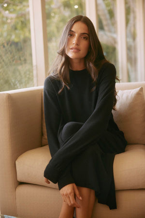A person with long hair in the AIDA DRESS by Velvet by Graham & Spencer, featuring a detachable belt, sits on a beige sofa by a large window.