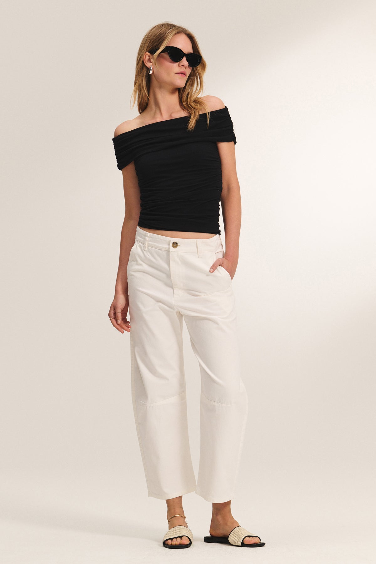 A woman stands against a plain background wearing the Velvet by Graham & Spencer ALEXANDRA TEE in black, styled with white wide-leg pants, black sunglasses, and sandals.-39149180715201