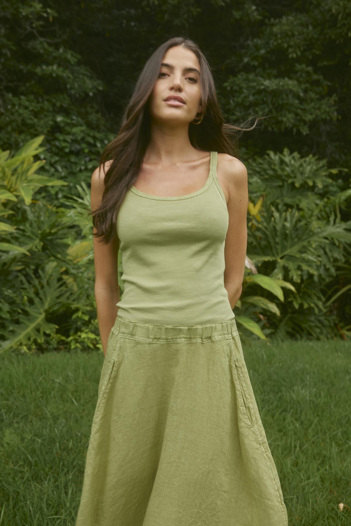 A woman stands in a lush outdoor setting wearing a light green tank top and an A-line skirt with an elastic waistband, the FAE LINEN SKIRT by Velvet by Graham & Spencer, both made from spring-weight linen.