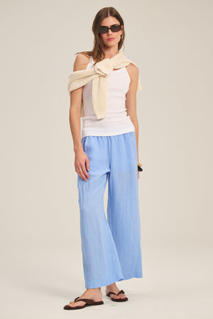 A woman wears LOLA LINEN PANT by Velvet by Graham & Spencer with a white tank top, cream sweater over her shoulders, black sunglasses, and brown sandals, standing against a plain background.