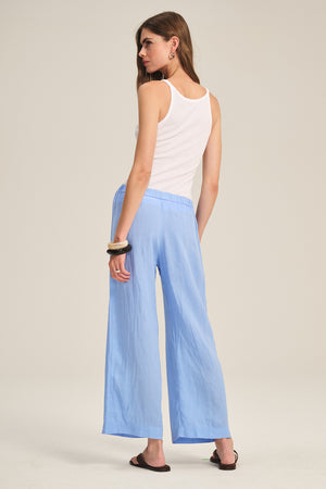 A person stands facing away, wearing a white sleeveless top, black sandals, hoop earrings, a bangle, and Velvet by Graham & Spencer's LOLA LINEN PANT—light blue with an elastic waist and wide-leg style.