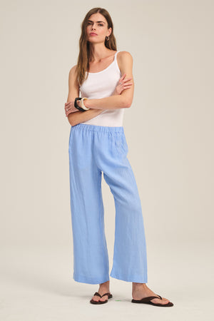 A woman stands against a plain background wearing a white tank top, brown sandals, and the LOLA LINEN PANT in light blue by Velvet by Graham & Spencer, featuring an elastic waist and pull-on style.
