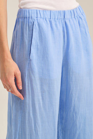 A person wears the LOLA LINEN PANT by Velvet by Graham & Spencer—light blue, lightweight linen trousers with an elastic waist—paired with a white top, their left hand resting at their side.