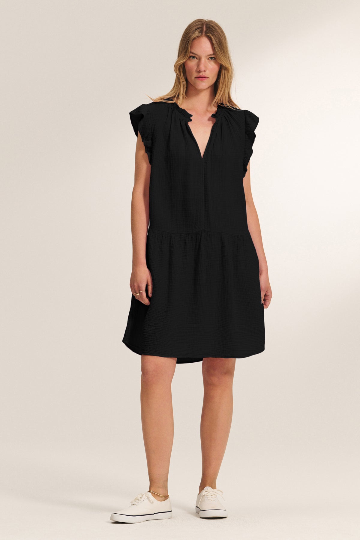 A person faces forward wearing the ANNIE COTTON GAUZE DRESS by Velvet by Graham & Spencer—a black, short-sleeve, knee-length dress with a V-neckline—paired with white sneakers against a plain background.-39172969103553