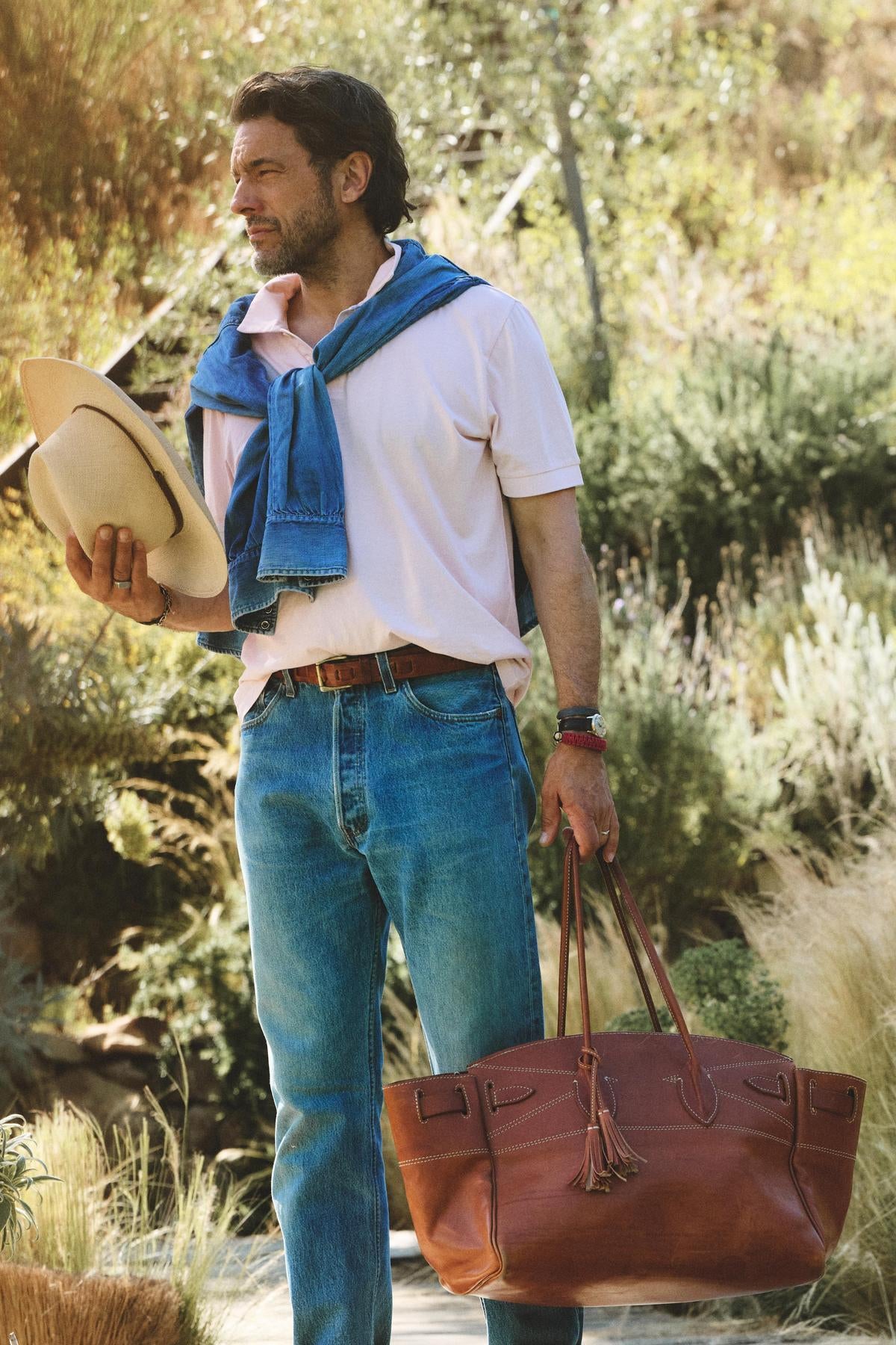 Wearing the ARROW POLO by Velvet by Graham & Spencer with jeans and a blue sweater over his shoulders, a man stands outdoors holding a hat and a brown leather bag.-41017028378817