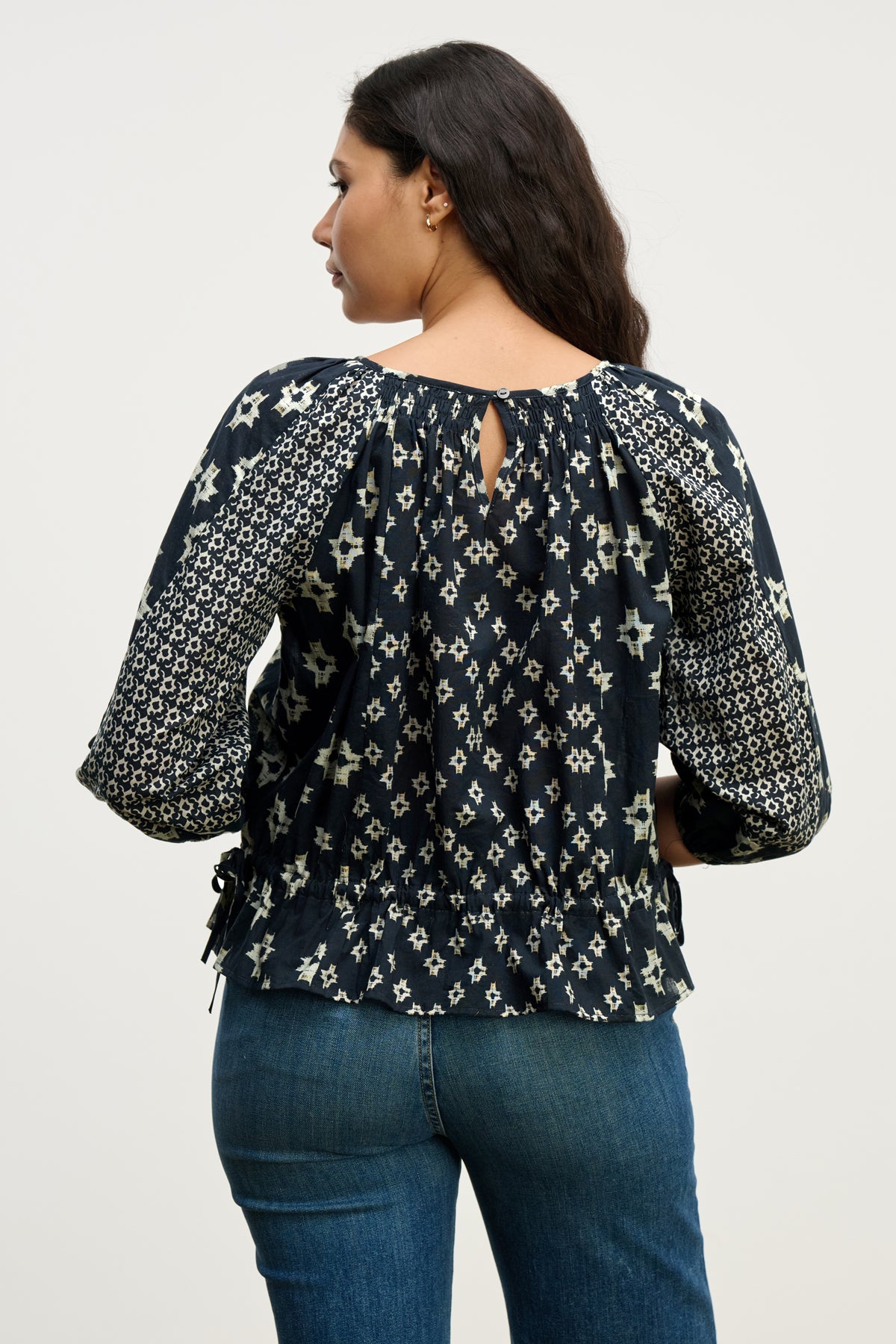 A woman with long dark hair is seen from behind, wearing the Velvet by Graham & Spencer ADELAIDE TOP in navy blue with ¾ sleeves and blue jeans against a plain background.-40227019653313